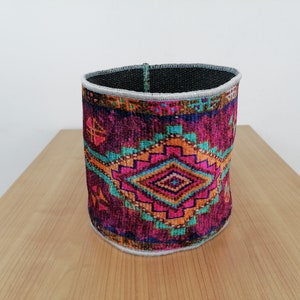 May include: A cylindrical fabric storage basket with a colorful geometric pattern in shades of pink, orange, teal, and purple. The basket has a black fabric lining and a white trim around the top.