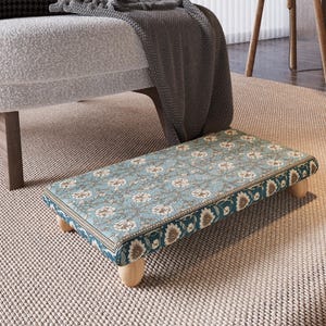 May include: A rectangular footstool with a floral pattern in shades of blue, white, and brown. The footstool has a dark teal border and is supported by four short, light-colored wooden legs. It is placed on a neutral-colored rug.