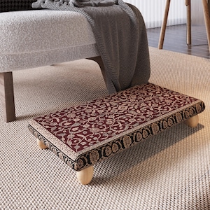 Puede incluir: Un reposapiés rectangular con un estampado floral burdeos y un borde negro con detalles dorados. Se apoya sobre cuatro patas cortas de madera clara. El reposapiés está colocado sobre una alfombra de color neutro.