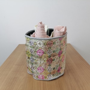 May include: A cylindrical storage basket with a floral pattern in shades of pink, green, and yellow. The basket is made of fabric and has a silver trim. It is filled with folded towels.