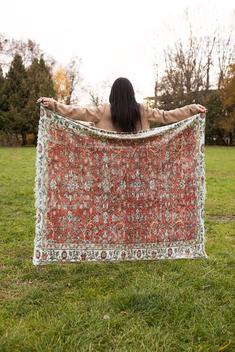 May include: A woman holds a large, red and white patterned rug in front of her. The rug has a floral design and a fringed edge. The woman is standing in a grassy field with trees in the background.