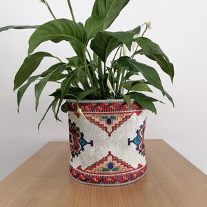 Puede incluir: Una planta de lirio de la paz verde en una maceta blanca con un patrón geométrico colorido. La maceta tiene un diseño rojo, naranja, azul y verde.
