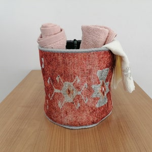 May include: A red and gray patterned fabric storage basket with a rolled up pink towel and a white towel inside.