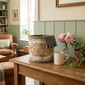 May include: A cylindrical storage basket featuring a white and gold ornate damask pattern with a dark brown woven trim. A folded grey and white herringbone patterned textile is draped over the rim of the basket, which sits on a wooden table.