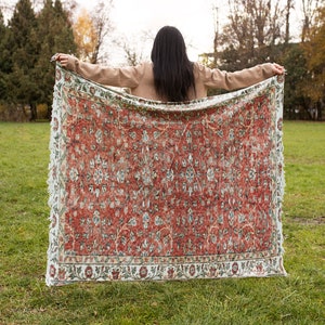 May include: A woman holds a large, red and white patterned rug in front of her. The rug has a floral design and a fringed edge. The woman is standing in a grassy field with trees in the background.