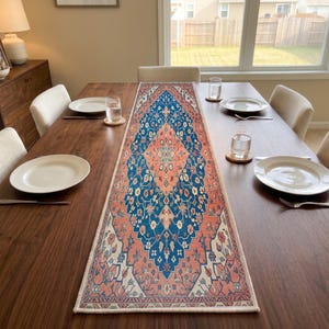 May include: A rectangular table runner featuring a central diamond-shaped medallion pattern in shades of deep blue, coral pink, and cream. The intricate floral and geometric motifs are printed across the fabric, which is laid flat on a wooden table.
