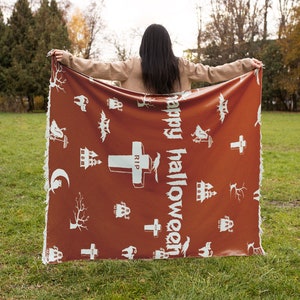 Op de afbeelding: Een persoon houdt een bruine deken vast met een witte tekst "Happy Halloween" en verschillende Halloween-thema illustraties, waaronder geesten, vleermuizen, kruisen en spookhuizen.