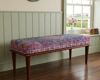 May include: A rectangular bench featuring a cushioned seat upholstered in a purple and blue fabric with an intricate, ornate pattern. The bench is supported by four tapered wooden legs in a medium brown finish, providing a stable base.
