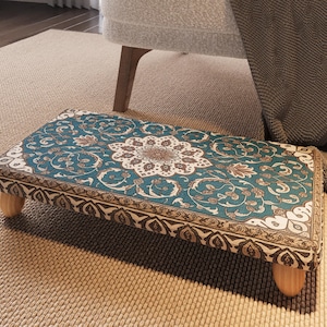 May include: A rectangular footstool with a teal and beige floral pattern. The footstool has a wooden frame and four short, rounded wooden legs. The design features a central medallion and ornate border.