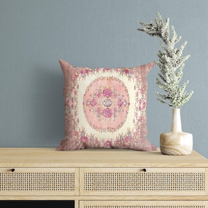 May include: A square throw cushion featuring a central circular medallion with pink floral patterns on a cream background, surrounded by a decorative border of pink and purple flowers and a textured, dusty rose-coloured outer edge.