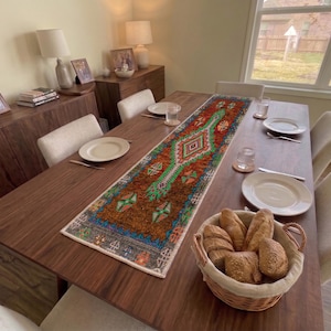 Op de afbeelding: Een rechthoekige tafelloper met een levendig, veelkleurig geometrisch patroon in tinten oranje, diepblauw en felgroen, geplaatst op een donkerhouten eettafel met witte borden, glazen en een mandje met broodjes.