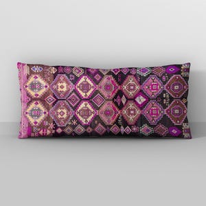Könnte beinhalten: Ein rechteckiges Lendenkissen mit dunklem Hintergrund und einem sich wiederholenden geometrischen Muster in Magenta-, Lila-, Braun- und Cremetönen. Das Design umfasst komplexe Rautenformen und kleine Motive, wobei der Text SALIHA sichtbar ist.