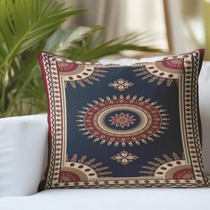May include: Decorative square pillow with a dark blue background and an intricate pattern in red, beige, and gold. The design features a central oval with a floral motif and sunburst-like elements. The pillow is on a white sofa.