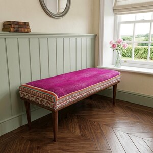 a cozy corner with a pink bench, a wooden shelf with books, and a window with a vase of pink flowers.