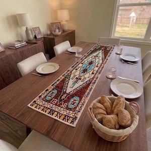 Op de afbeelding: Een rechthoekige tafelloper met een centraal geometrisch ruitpatroon in de kleuren rood, blauw, wit en geel. De loper ligt in de lengte in het midden van een donkerhouten eettafel, die is gedekt met witte borden en glazen.