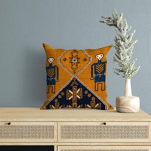 May include: A square throw pillow featuring a mustard yellow and dark blue geometric pattern with stylized human figures and diamond motifs. The design includes white and black linear details, creating a symmetrical composition across the fabric.