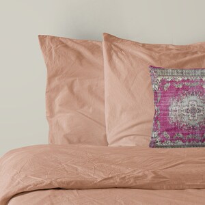 May include: A pink and gray patterned throw pillow with a decorative border. The pillow is on a peach-colored bed with two matching pillows.