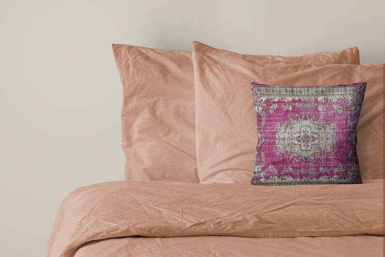May include: A pink and gray patterned throw pillow with a decorative border. The pillow is on a peach-colored bed with two matching pillows.