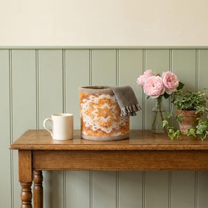 Peut inclure: Un panier de rangement cylindrique en tissu avec un motif géométrique orange et blanc et une bordure à chevrons gris foncé. À côté se trouvent une tasse en céramique blanche, un vase en verre avec trois roses roses et une plante lierre verte.