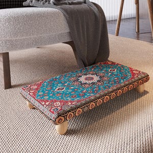 May include: A rectangular footstool with a vibrant, patterned fabric top in teal, red, and cream. The design features floral and geometric motifs. The stool is supported by four short, light-colored wooden legs.