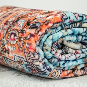 K&ouml;nnte beinhalten: Eine zusammengerollte Fleece-Decke mit einem farbenfrohen, komplizierten Muster. Die Decke hat eine Mischung aus orange, blau und wei&szlig;en Farben.