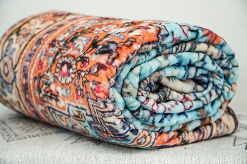 K&ouml;nnte beinhalten: Eine zusammengerollte Fleece-Decke mit einem farbenfrohen, komplizierten Muster. Die Decke hat eine Mischung aus orange, blau und wei&szlig;en Farben.