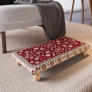 Könnte beinhalten: Ein rechteckiger Fußhocker mit rotem Blumenmuster und cremefarbenem Rand. Er wird von vier kurzen, hellen Holzbeinen getragen. Das Design weist einen traditionellen, verzierten Stil auf. Ca. 60 cm lang.