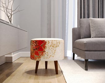 May include: A round ottoman with a white top and a patterned fabric skirt. The fabric features a red and gold floral design. The ottoman has three dark wooden legs and is placed on a hardwood floor.