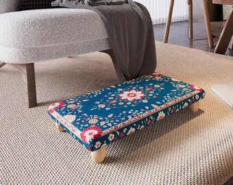 May include: A rectangular footstool with a teal floral pattern. The design features red and cream-colored flowers and foliage. The stool has four short, light-colored wooden legs and is placed on a neutral-colored rug.