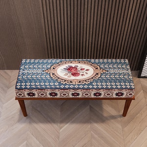 Puede incluir: Banco rectangular tapizado con diseño floral. El banco presenta un fondo azul con una pieza central floral ovalada en rojo y crema. Las patas de madera son de color marrón oscuro.