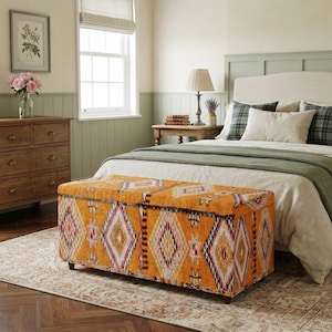 May include: A rectangular storage bench upholstered in a vibrant orange fabric featuring a repeating geometric diamond pattern in white, black, and pink. The bench is supported by short, dark brown wooden feet and has a flat, hinged top lid.