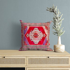 May include: A red, pink, and purple patterned throw pillow with a geometric design. The pillow is sitting on a wooden dresser with two drawers.