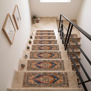 May include: A set of rectangular stair treads featuring a central diamond motif in shades of blue, orange, and cream. The treads are placed on light beige stone stairs with a black metal railing and small potted succulents on the side.