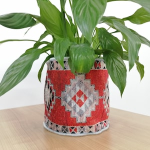 May include: A green plant in a red and white patterned fabric planter. The planter has a geometric design with black and white accents.