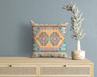 Puede incluir: Un cojín cuadrado con un panel central de color melocotón que presenta dos motivos geométricos simétricos en tonos rosa, azul cerceta y negro. El diseño está enmarcado por un borde decorativo con patrones intrincados en azul, blanco y negro.