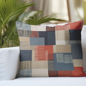 Puede incluir: Cojín cuadrado con diseño de patchwork. El cojín presenta un patrón geométrico de rectángulos en tonos rojos, azules, beige y negro. Está colocado sobre un sofá blanco, con una planta verde al fondo.
