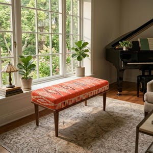 May include: A rectangular bench featuring a vibrant orange fabric seat with a white and black geometric diamond pattern. The bench is supported by four tapered wooden legs in a medium brown finish, providing a stable base for the upholstered top.