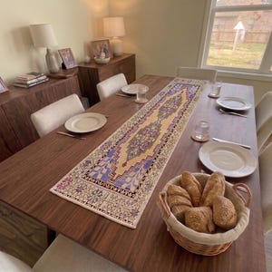 Op de afbeelding: Een rechthoekige tafelloper met een centraal medaillonpatroon in blauwe, paarse en crèmetinten, plat op een donkerhouten eettafel gelegd. De loper heeft een gedetailleerde geometrische rand en ligt tussen witte borden en glazen in.