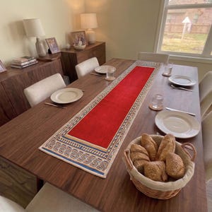 Puede incluir: Un camino de mesa rectangular con un panel central de color rojo vibrante y un borde decorativo con patrones geométricos en azul, crema y marrón. El camino está colocado a lo largo sobre una mesa de comedor de madera oscura con platos blancos.