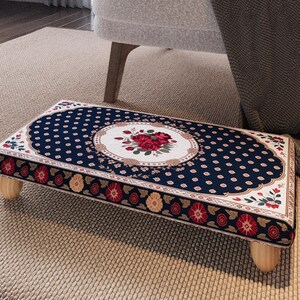 Peut inclure: Un repose-pieds rectangulaire avec un dessus bleu marine orné d'un motif floral. Le centre présente un ovale blanc avec des roses rouges. Les bords sont ornés de motifs floraux rouges et jaunes. Il repose sur quatre pieds en bois clairs.