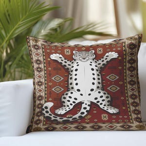 May include: Square throw pillow featuring a cartoon snow leopard with black spots, arms outstretched, on a red patterned rug design. The pillow is on a white couch with a brown pillow to the right. Decorative home decor.