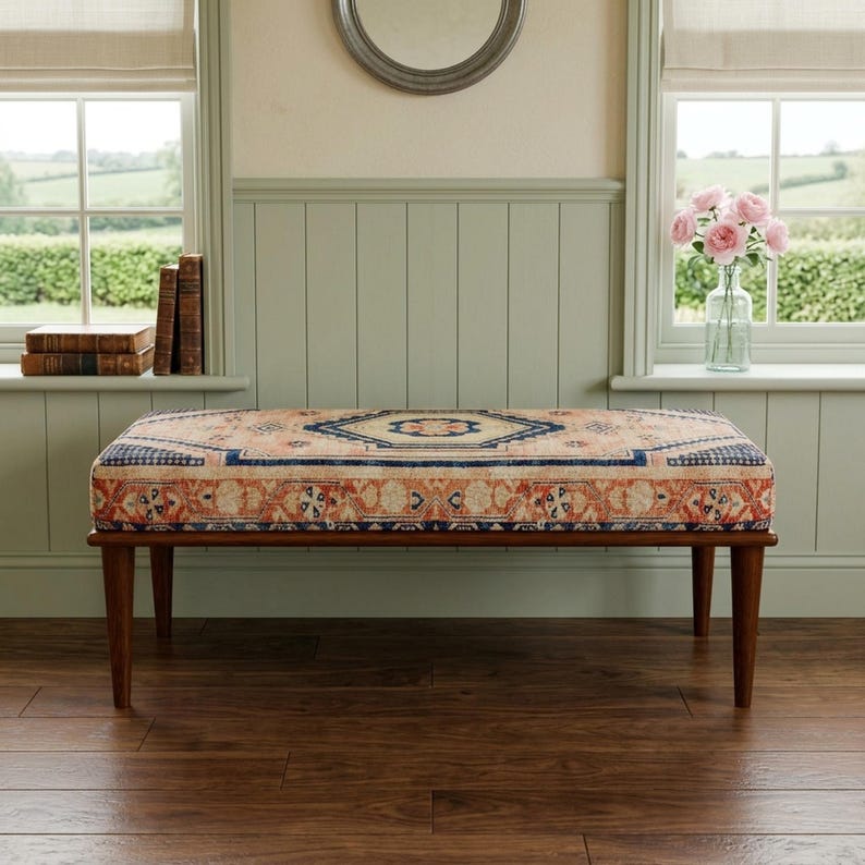a wooden bench with a colorful, patterned seat, placed in front of a window with a vase of pink flowers on the sill.