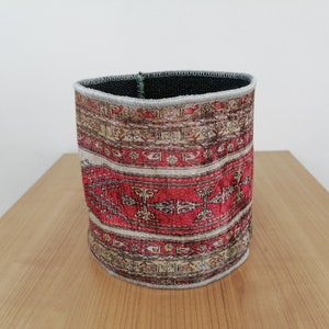 Puede incluir: Contenedor cilíndrico con un diseño estampado que recuerda a una alfombra tradicional. El contenedor presenta una combinación de colores rojo, beige y marrón con intrincados patrones geométricos. El borde está acabado con un ribete gris.