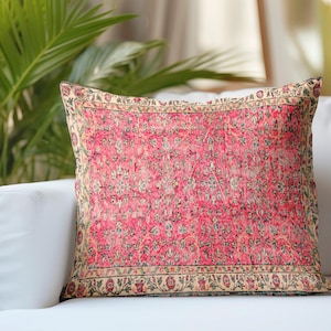 Puede incluir: Un cojín cuadrado con un centro rosa vibrante y un patrón detallado de flores y enredaderas en tonos verde, crema y rojo oscuro. El cojín está enmarcado por un borde decorativo con un motivo botánico repetitivo en colores a juego.