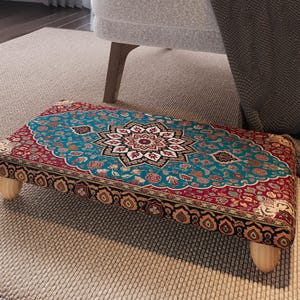May include: A rectangular footstool with a detailed, colorful rug design. The rug features a central medallion in shades of teal, red, and cream, surrounded by floral patterns. The footstool is supported by four short, light-colored wooden legs.