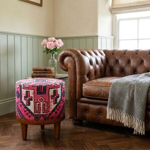 a cozy living room with a brown leather sofa, a round ottoman with a colorful pattern, a vase of pink roses, and a stack of books on a small side table.