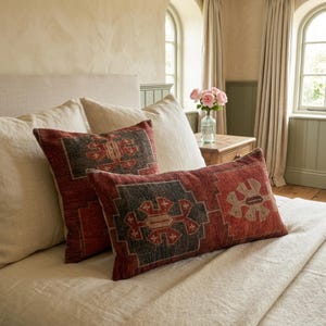 a cozy bedroom with a bed adorned with two decorative pillows, one with a red and black pattern and the other with a floral design. A vase of pink flowers is placed on a nightstand next to the bed, adding a touch of elegance to the room.