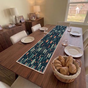 May include: A rectangular table runner featuring a dark teal background with a repeating pattern of stylized orange, light blue, and white vertical shapes. The runner has a light-colored stitched border and is centered on a dark wood dining table.