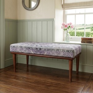 a wooden bench with a purple and white patterned cushion, placed on a wooden floor in a room with a window and a bookshelf.