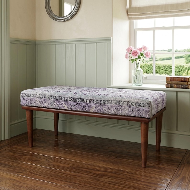 a wooden bench with a purple and white patterned cushion, placed on a wooden floor in a room with a window and a bookshelf.